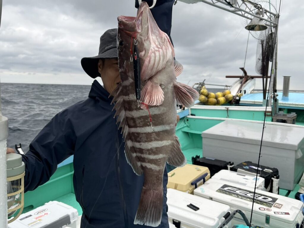 黒いジャケットと帽子をかぶった人物が、船の上で大きな縞模様の魚を誇らしげに掲げています。