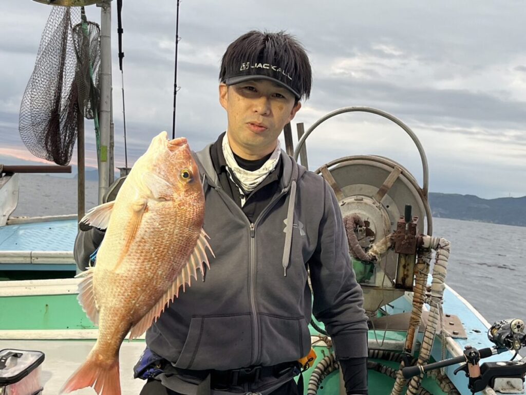 釣った大きな鯛を抱えたボートの上の人物。黒いジャケット、ヘッドバンド、手袋を着用している。