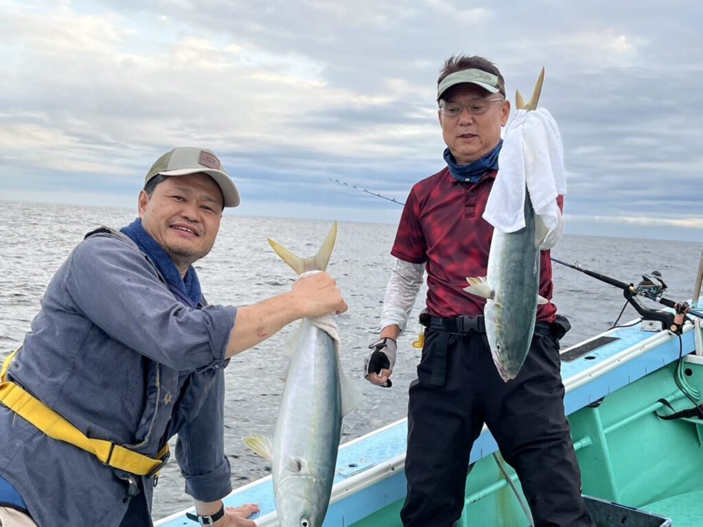 2 人が獲れたての魚を抱えています。2 人は笑顔で、1 人は赤いシャツと眼鏡をかけ、もう 1 人は青いジャケットと帽子をかぶっています。背景には海と曇り空が見えます。