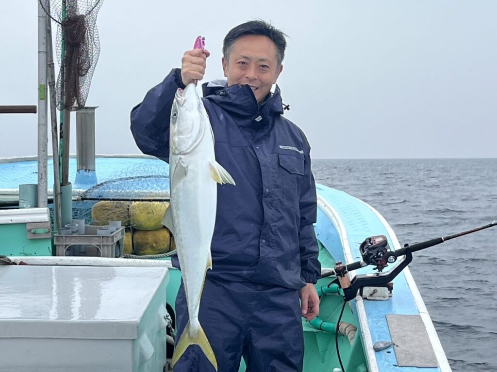 遊漁船に乗って、釣った大きな魚を抱えている人。青いジャケットを着て、釣り道具の横に立っています。