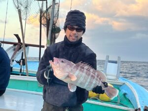 黒い服とサングラスをかけた人物が、海と空を背景に、ボートの上で大きな魚を抱えている。ボートには漁網が見える。.