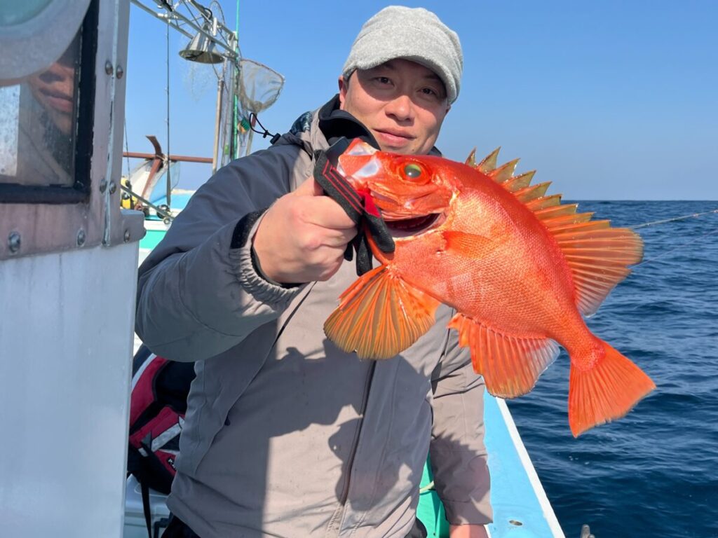 とがったひれを持つ鮮やかなオレンジ色の魚を抱えたボートの上の人物。背景の空は晴れ、水面は穏やかです。人物は灰色のジャケットと帽子をかぶっています。.