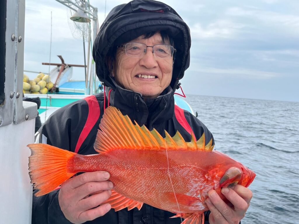 黒いジャケットとフードを身に着け、笑顔で魚をボートの上で抱えている人物。