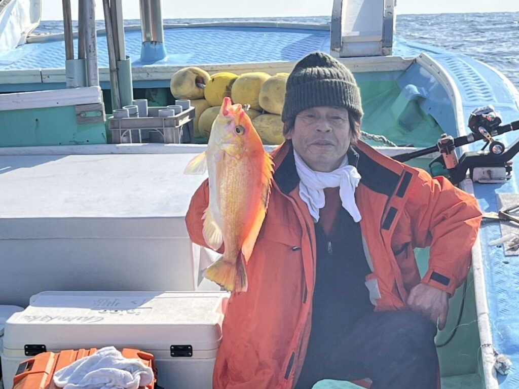 オレンジ色のジャケットと緑色のビーニー帽をかぶった人物がボートに乗り、大きな魚を抱えています。