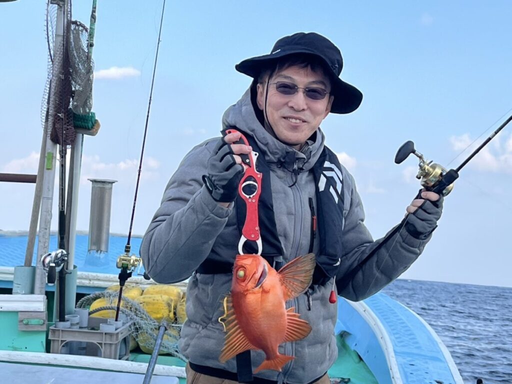 黒い帽子と灰色のジャケットを着た人物が、片手に釣り竿、もう片方の手には大きな魚を持ち、微笑んでいます。