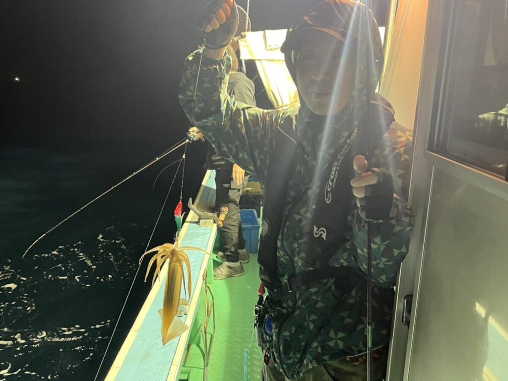 明るい光に照らされた夜、船の上に立ち、釣り糸につけたばかりのイカを掲げている。