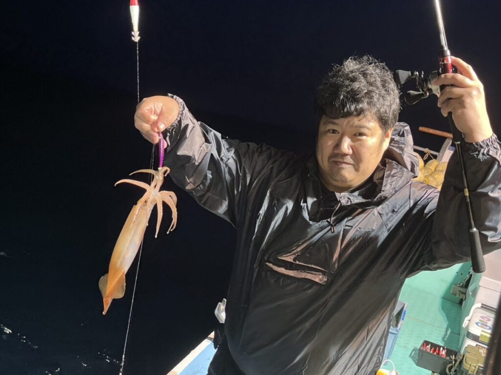 夜、レインジャケットを着た人が片手に釣り竿を持ち、もう片方の手には獲れたてのイカを持って船の上に立っています。