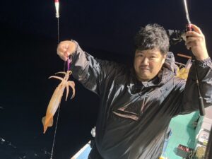 夜、レインジャケットを着た人が片手に釣り竿を持ち、もう片方の手には獲れたてのイカを持って船の上に立っています。.