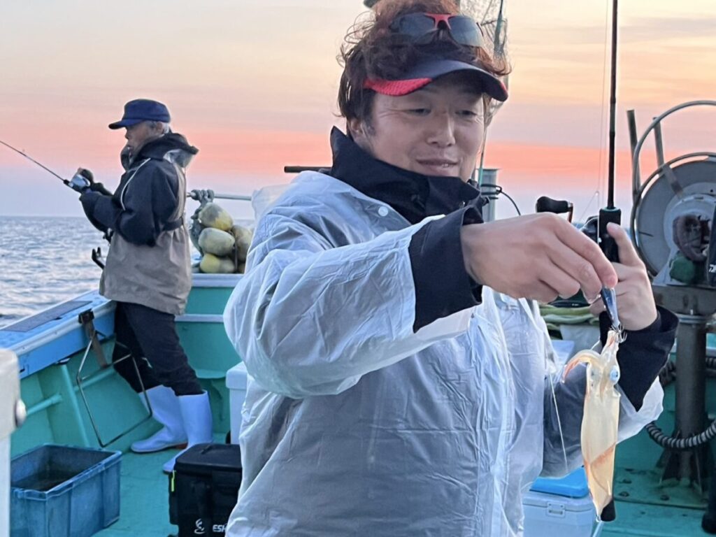 日没時のボート上で、レインジャケットを着た人が釣り糸につけたイカを持ちながら微笑んでいる。