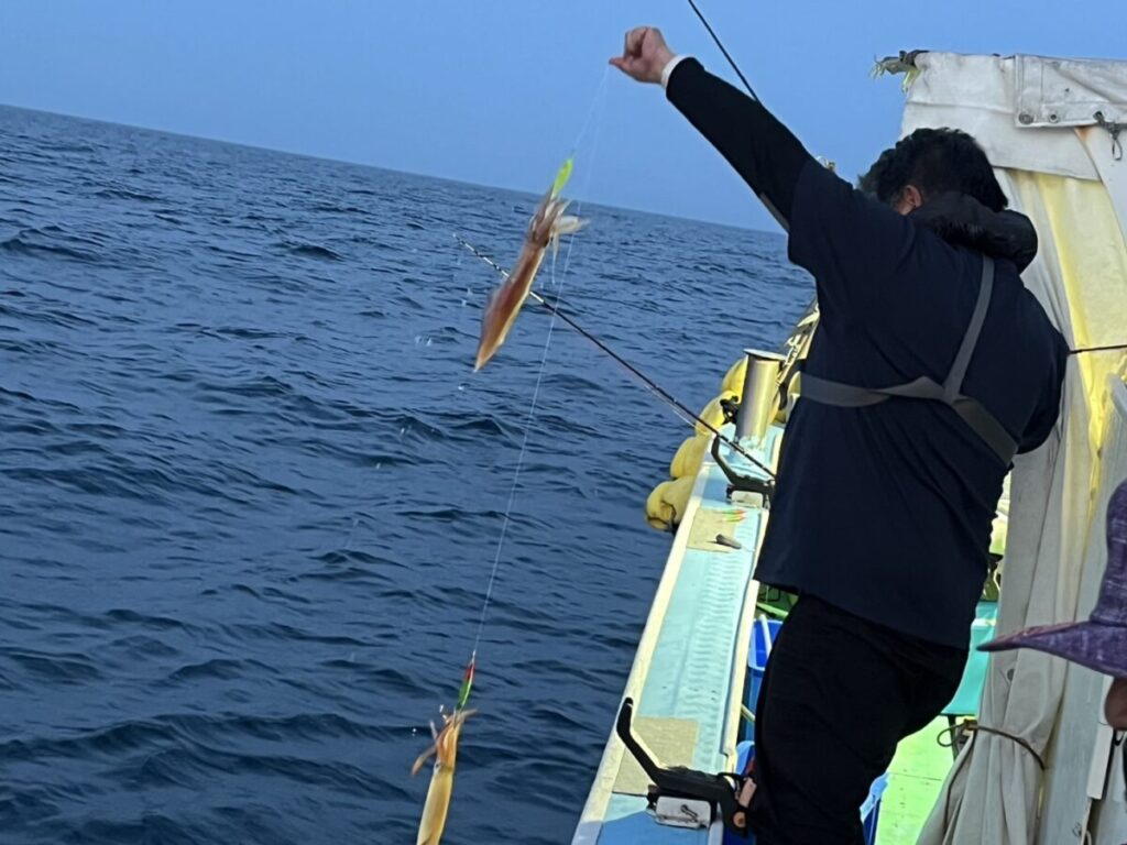 海と澄んだ空を背景に、船に乗った人が2匹のイカを釣り上げた釣り糸を引き上げています。