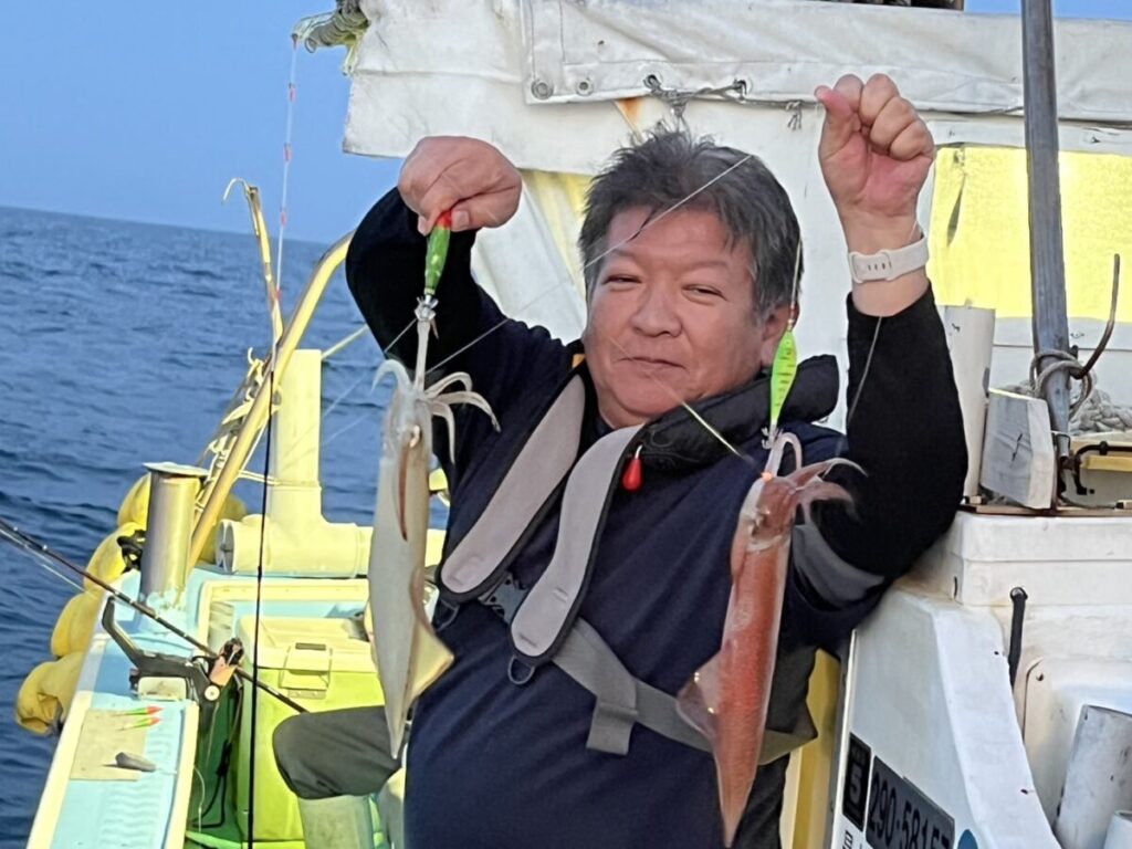 船に乗った男性が、両手に新鮮なイカを1匹ずつ持ち、微笑んでいる。ライフジャケットを着用し、周囲に漁具が見える海上にいる。.