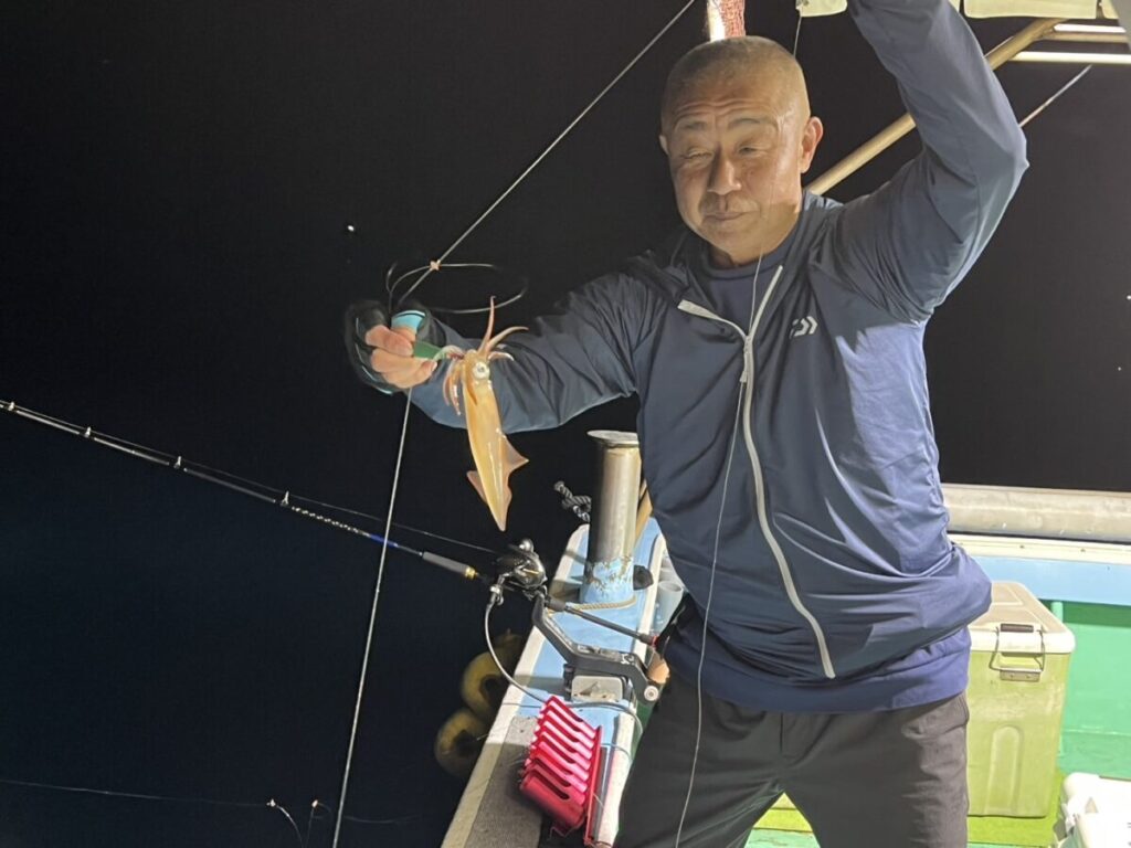 夜、船の上で釣り糸に釣ったばかりのイカを掲げた男性が、カメラに向かって微笑んでいる。背景は暗く、船はライトで照らされている。.