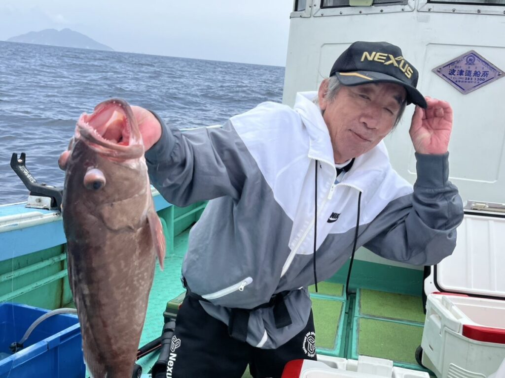 海上の船に乗った男性が、大きな魚を片手で持ち上げながら、微笑んで帽子を傾けている。