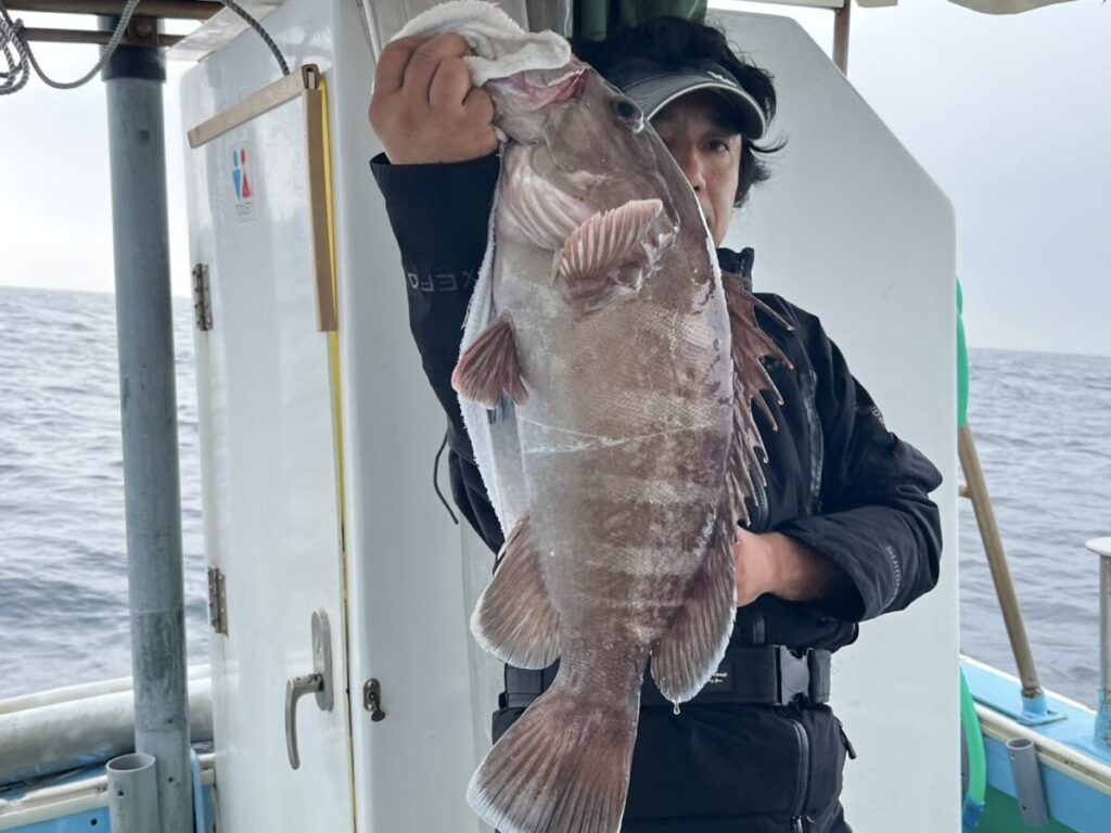 船に乗った人物が片手で大きな魚を持ち上げており、顔の一部が隠れている。背景には海と船内の一部が見える。