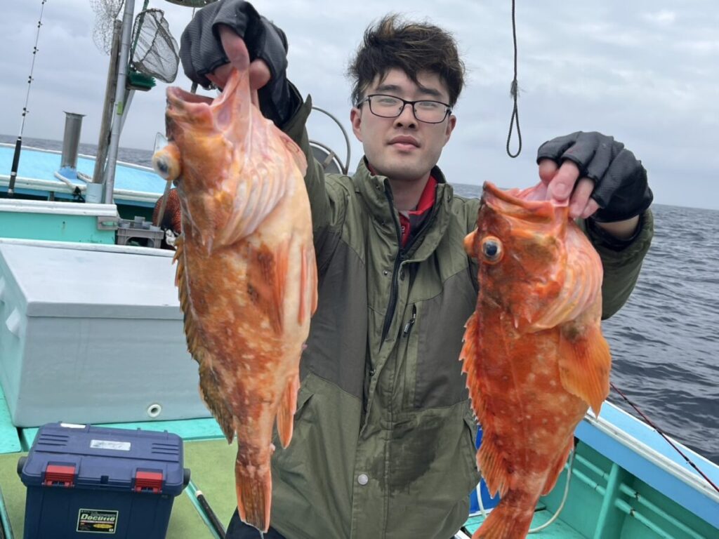 眼鏡と手袋をはめた人物が、海と曇り空を背景に、ボートの上で2匹の大きなオレンジ色の魚を持ち上げている。デッキには漁具とクーラーボックスが見える。.
