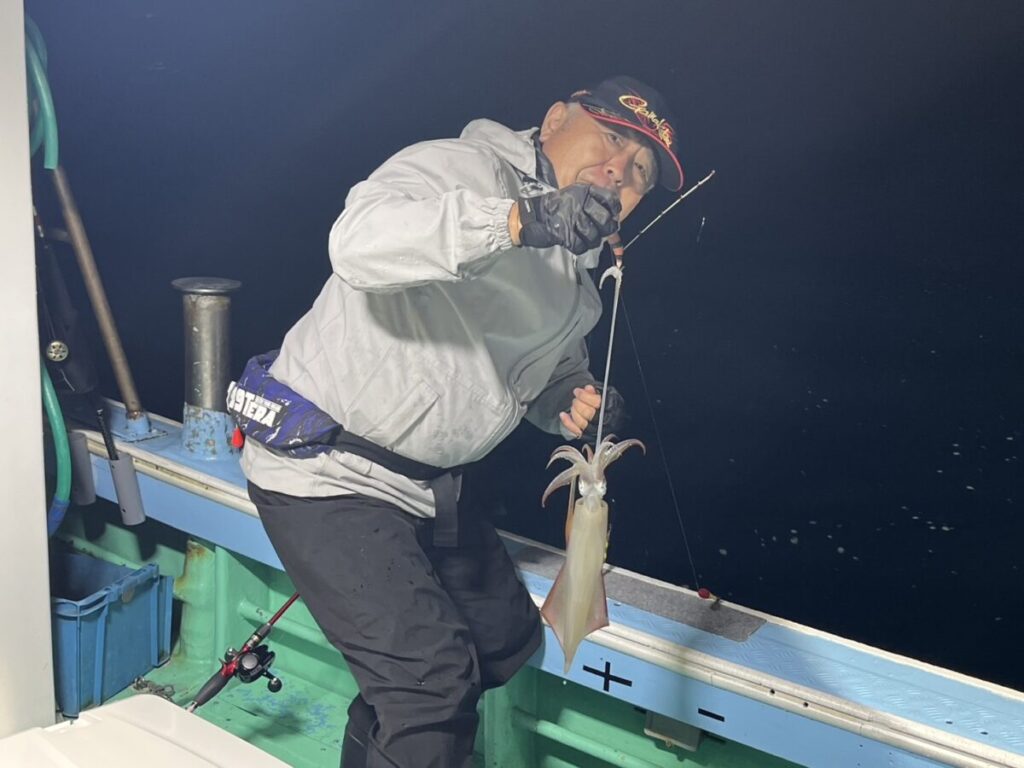 夜、帽子と手袋をはめた人物が船の上に立ち、釣り糸に釣ったばかりのイカを握っている。背景には船内と暗い水面が見える。