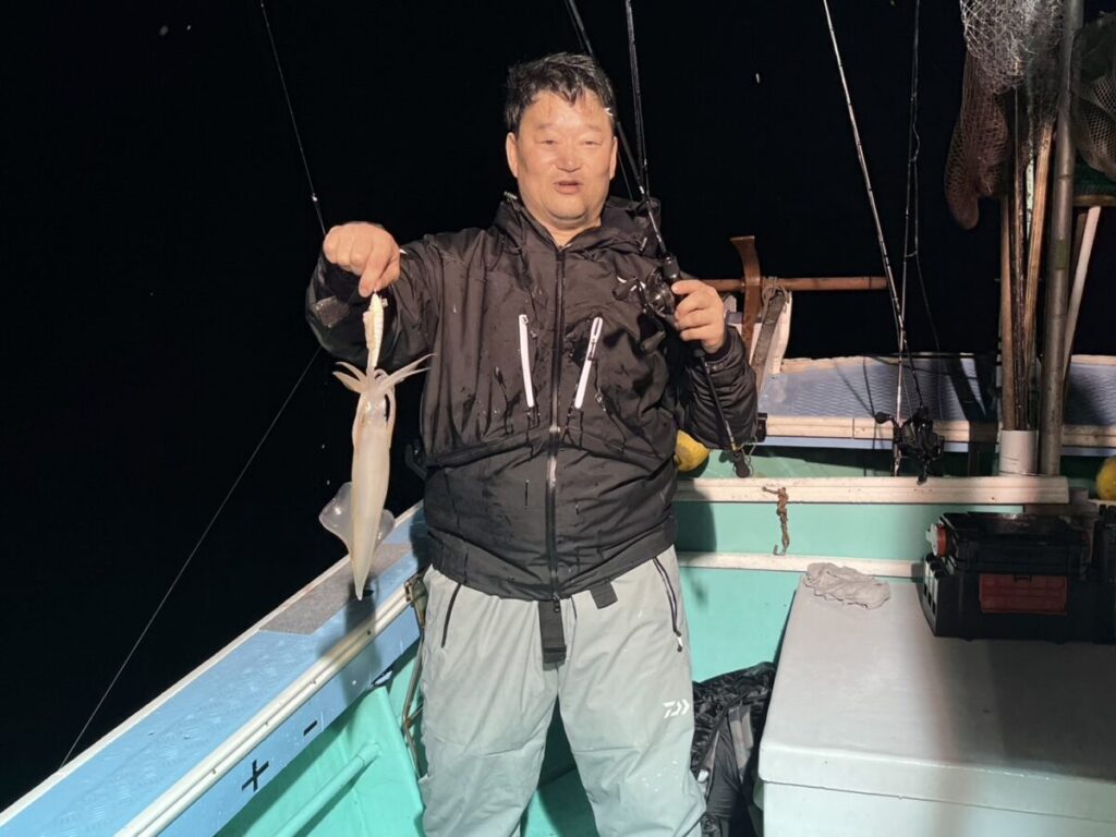 夜、船の上で、男性が釣り上げたばかりのイカを片手に持ち、カメラに向かって微笑んでいる。黒いジャケットとライトグレーのパンツを羽織っている。周囲にはデッキと漁具が見える。.