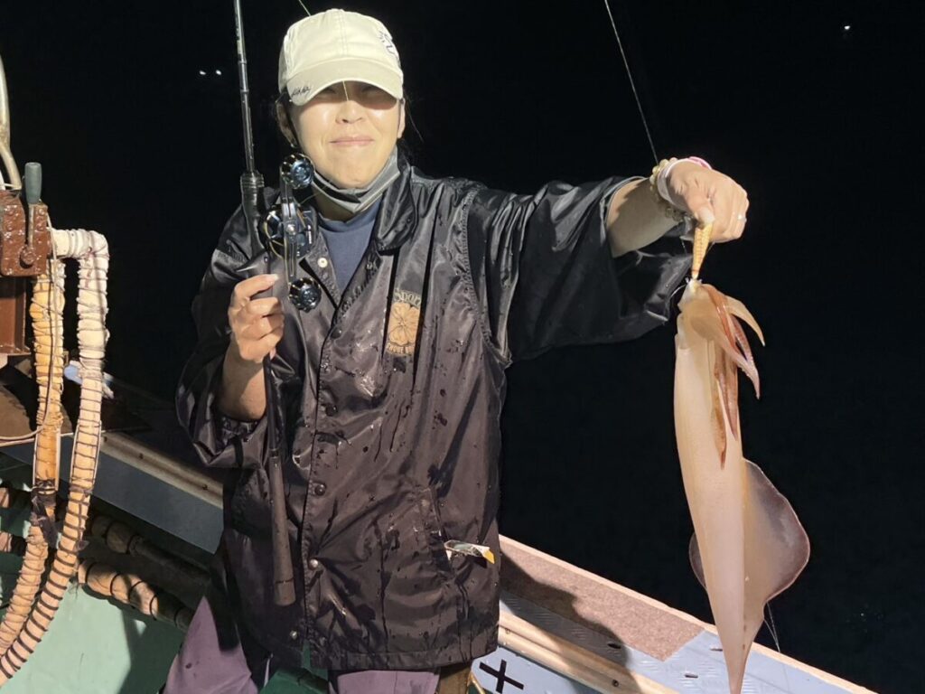 明るい色の帽子と黒いジャケットを着た人物が夜、ボートの上に立ち、片手に釣り竿を持ち、もう片方の手には獲れたての大きなイカを見せながら、カメラに向かって微笑んでいる。.