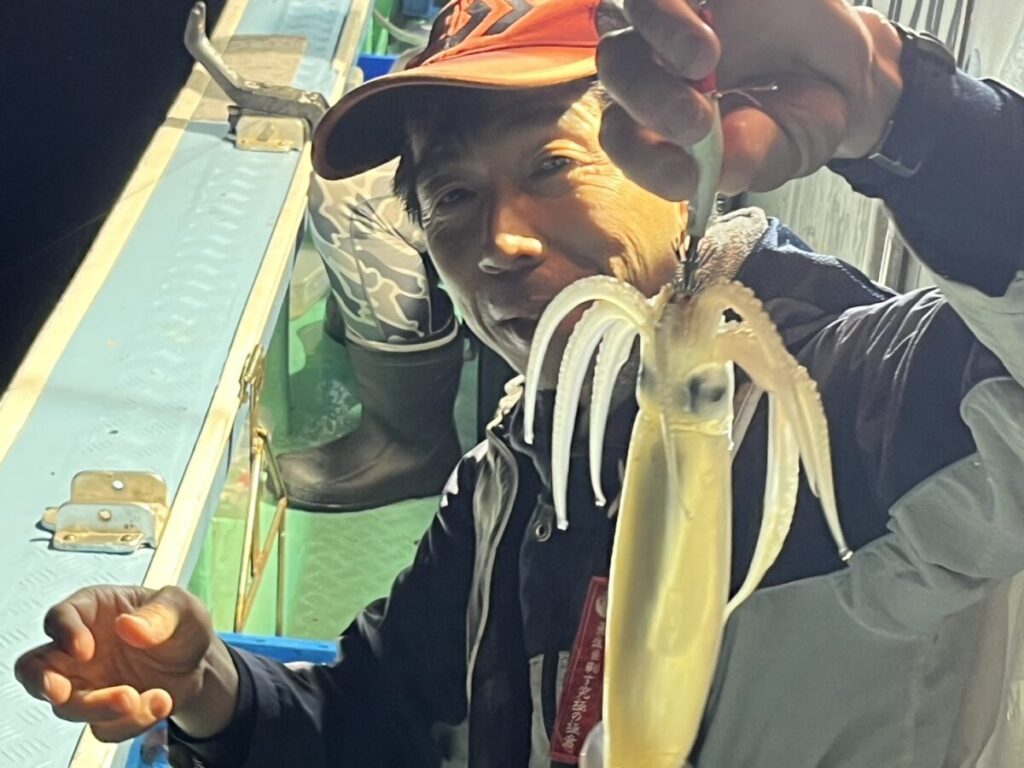 船の上で、釣り道具を使って獲れたてのイカを掲げる人物。夜の風景のようで、帽子とアウトドアウェアを身につけ、笑顔を浮かべている。.