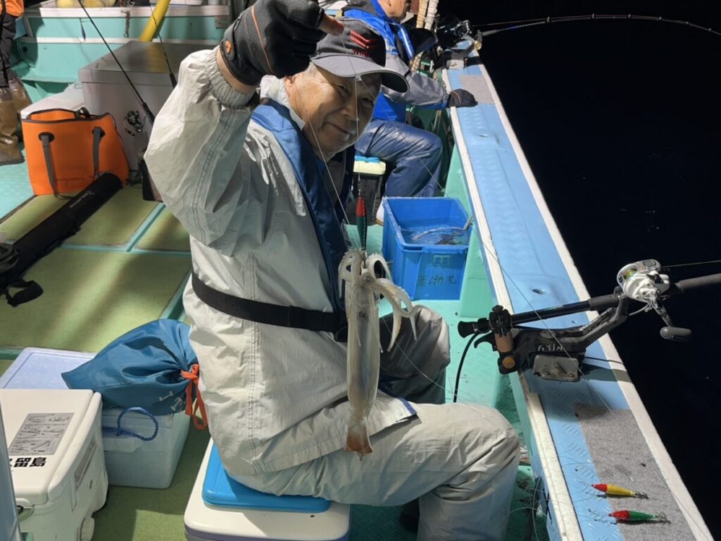 白い漁具、手袋、帽子を身に着けた年配の男性が、夜、船の上で釣り糸で釣ったイカを掲げながら微笑んでいる。周囲には漁具とコンテナが見える。