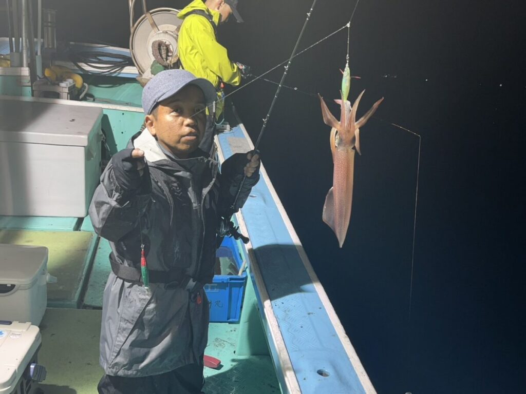 夜、ボートに乗っている人物が釣り竿を手に持ち、大きなイカが釣り糸にかかっている。黄色いジャケットを着た別の人物も近くに立ち、同じように釣りをしている。ボートのデッキは明るく照らされ、釣り道具が見える。.