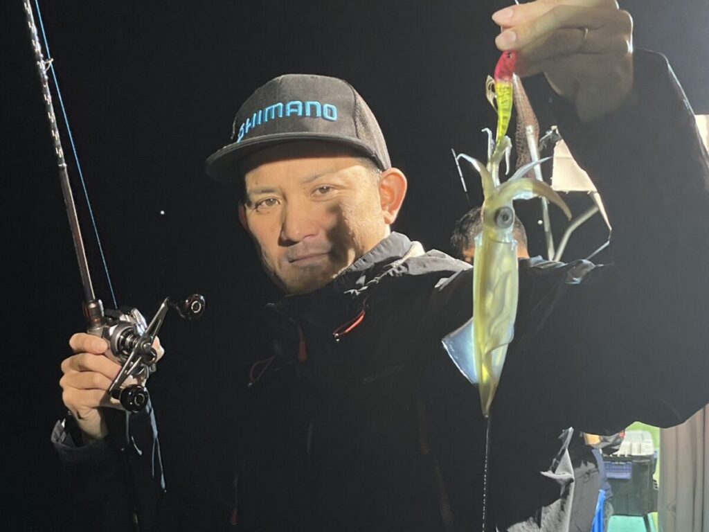 黒いシマノのキャップをかぶった男性が、夜に屋外に立って、片手に釣り竿を持ち、もう片方の手には釣ったばかりのイカを見せている。