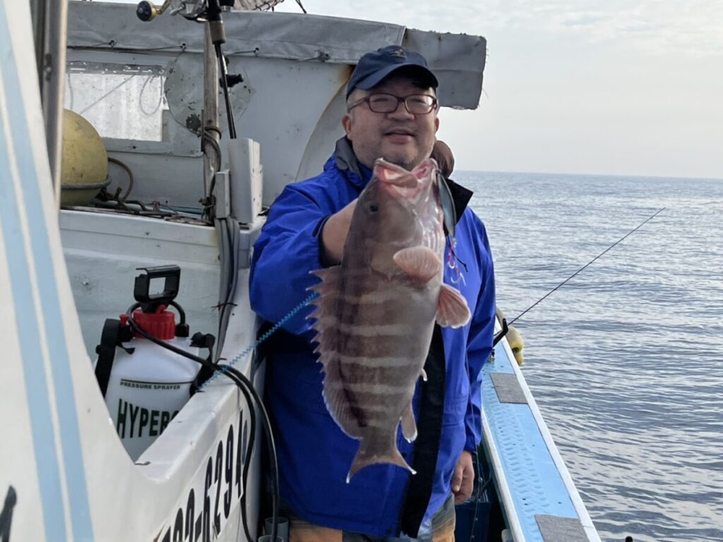 青いジャケットと帽子をかぶった人物が船の上に立ち、笑顔で大きな縞模様の魚を掲げています。背景には海と釣り竿が見えます。.