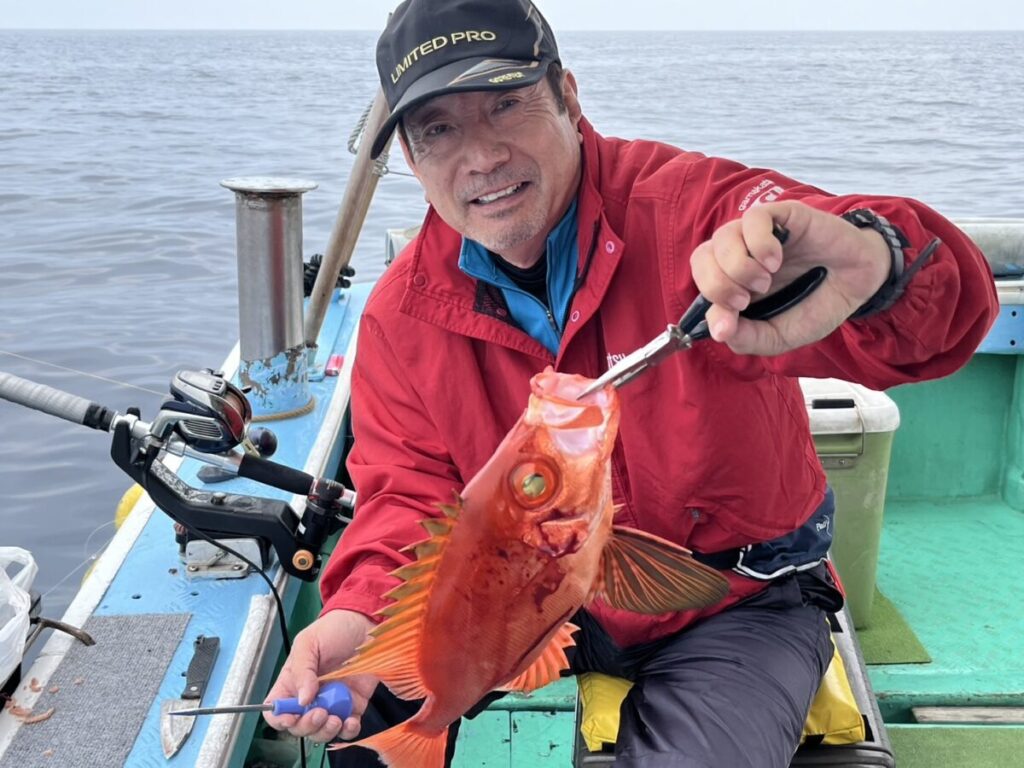 赤いジャケットと黒い帽子をかぶった男性が、沖合の漁船の上で、獲れたての鮮やかなオレンジ色の大きな目をした魚を抱えて微笑んでいる。周囲には漁具や漁具が見える。.