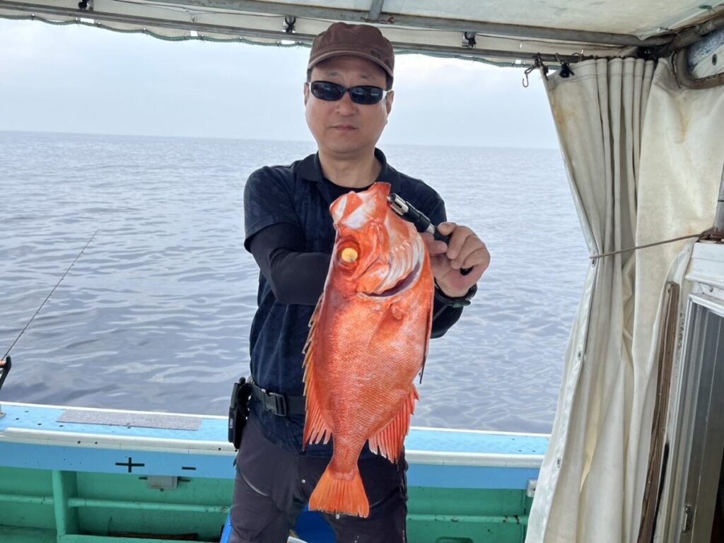 サングラスをかけ、茶色の帽子をかぶった人物が、海を背景にしたボートの上で、大きくて鮮やかな赤い魚を抱えています。.