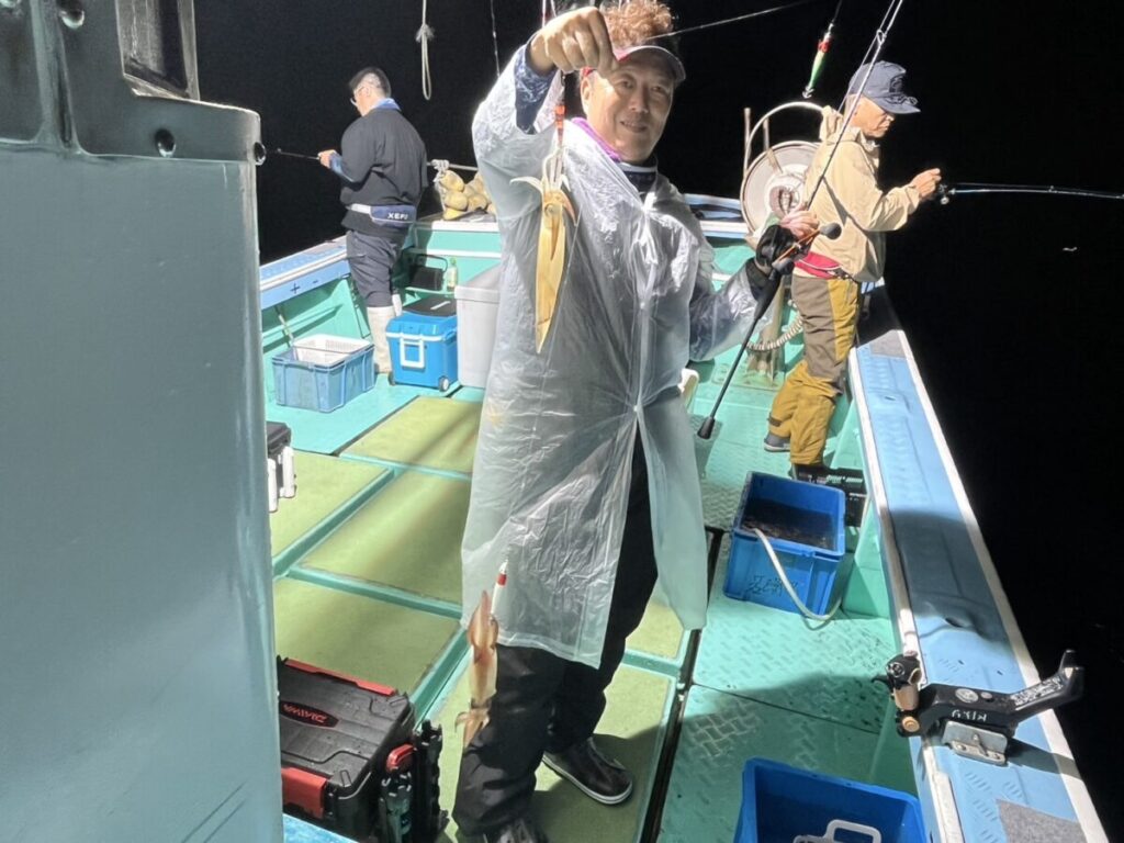 夜、漁船の上でレインポンチョを着た人が釣ったイカを掲げており、その後ろでは2人が釣りをしている。デッキには釣り道具とクーラーボックスが置かれている。.