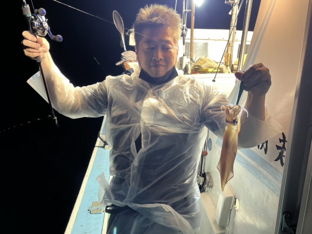 半透明のレインポンチョを着た人が、明るい夜のライトの下、船の上に立ち、片手に釣り竿、もう片方の手に獲れたてのイカを持っている。.