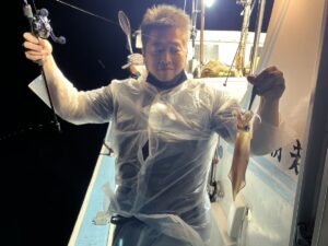 半透明のレインポンチョを着た人が、明るい夜のライトの下、船の上に立ち、片手に釣り竿、もう片方の手に獲れたてのイカを持っている。.