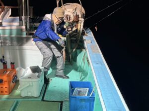 青いジャケットと帽子をかぶった人物が、船の上で夜釣りをしている。デッキの青い容器の上に釣り竿と釣ったイカを抱えている。その光景は人工照明で照らされている。.
