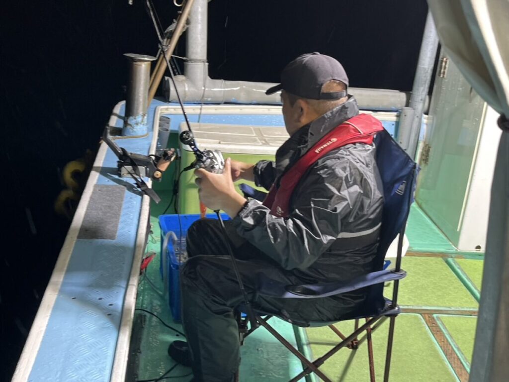 夜、黒い帽子、レインジャケット、赤いライフジャケットを着た人物が、ボートの上で折りたたみ椅子に座り、釣り竿とリールを手に釣りの準備をしている。ボートのデッキは濡れており、近くには釣り道具が置かれている。