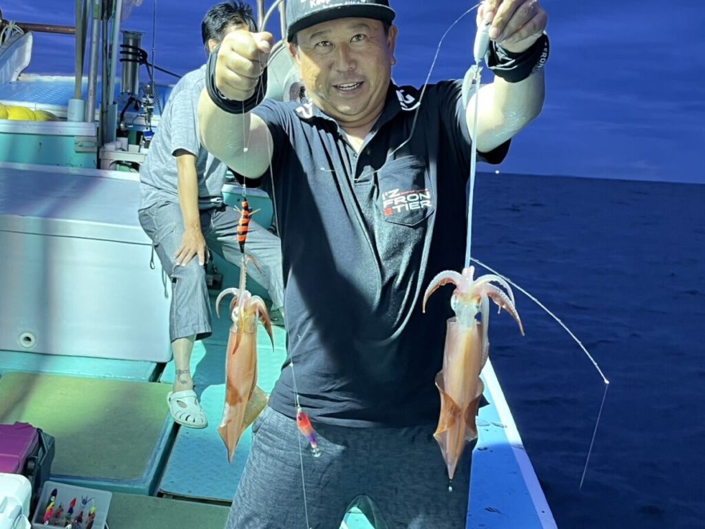 ボートに乗った男が、獲れたてのイカを両手に2杯ずつ掲げ、カメラに向かって微笑んでいる。彼の後ろにはもう一人座り、背景には青空の下に海が見える。.