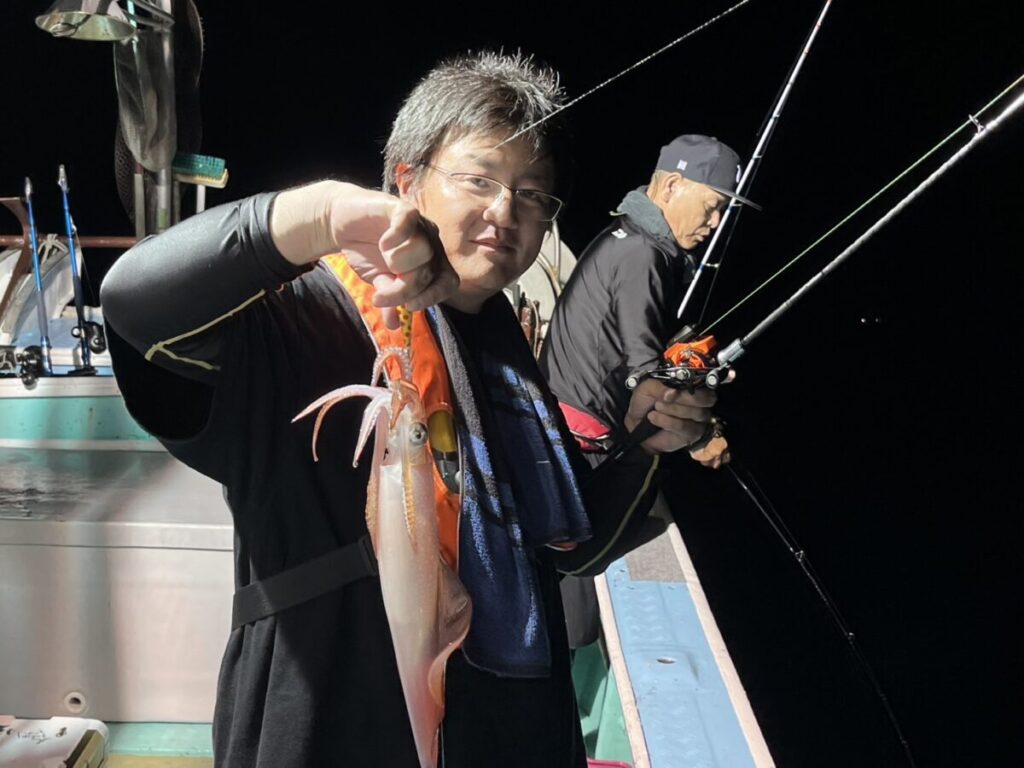 夜の船上で、釣りたてのイカを誇らしげに掲げる人と、その横で竿を持って釣りをしている人がいる。明るい照明の下、2人とも集中している。