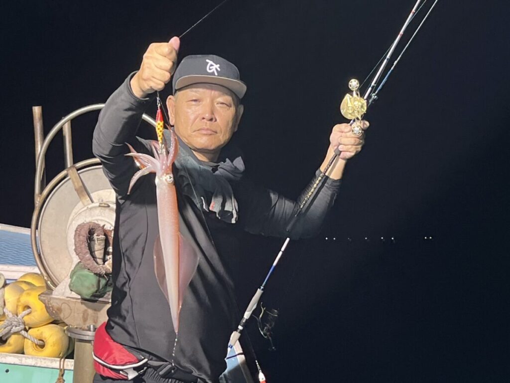 夜の船上で、釣り上げたばかりの大きなイカを釣り竿に掲げ、誇らしげに見せている人物。背景は暗く、漁具やかすかな明かりが見える。.