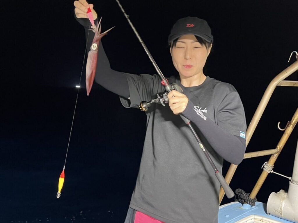 黒いシャツに帽子をかぶった人物が、イカがかかった釣り竿を持ち、暗い海を背景に夜の船の上に立っている。
