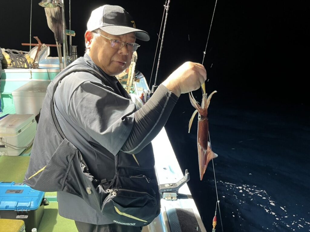 夜の船上で、釣り糸を垂れた釣りたてのイカを掲げる人。背景には釣り道具とタックルボックスが見える。