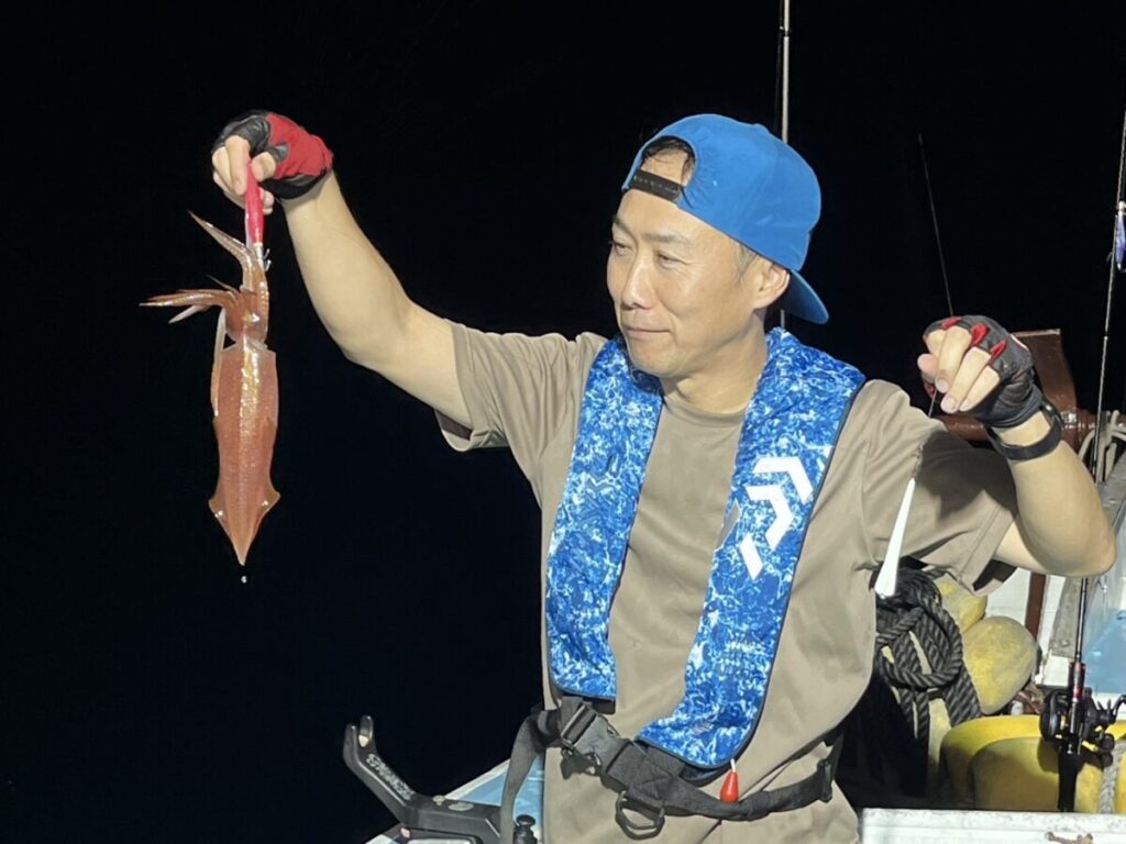 青い帽子に青い柄の救命胴衣、手袋をはめた男が、夜の船上で釣りたてのイカを掲げている。背景は暗く、釣果と釣り人を際立たせている。