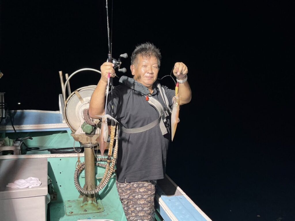 夜のボートの上で、片手に釣り竿、片手に釣りたてのイカを持ち、明るい光の下でカメラに向かって微笑む男。