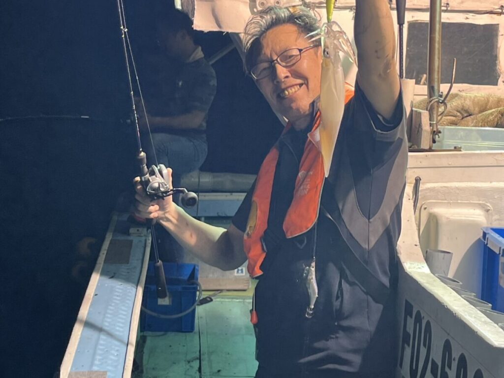 眼鏡をかけ、オレンジ色の救命胴衣を着た笑顔の人物が、夜のボートの上でイカがかかった釣り竿を掲げている。