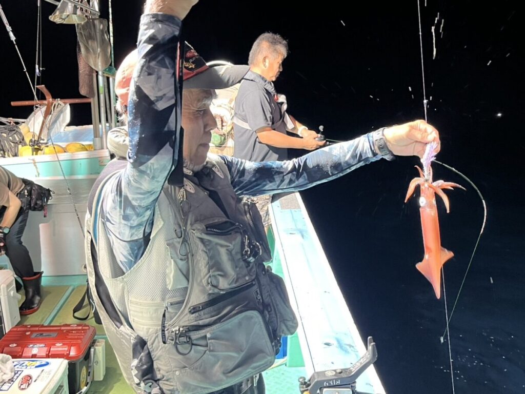 夜の船上で釣りベストを着た男が釣りたての赤イカを掲げる。
