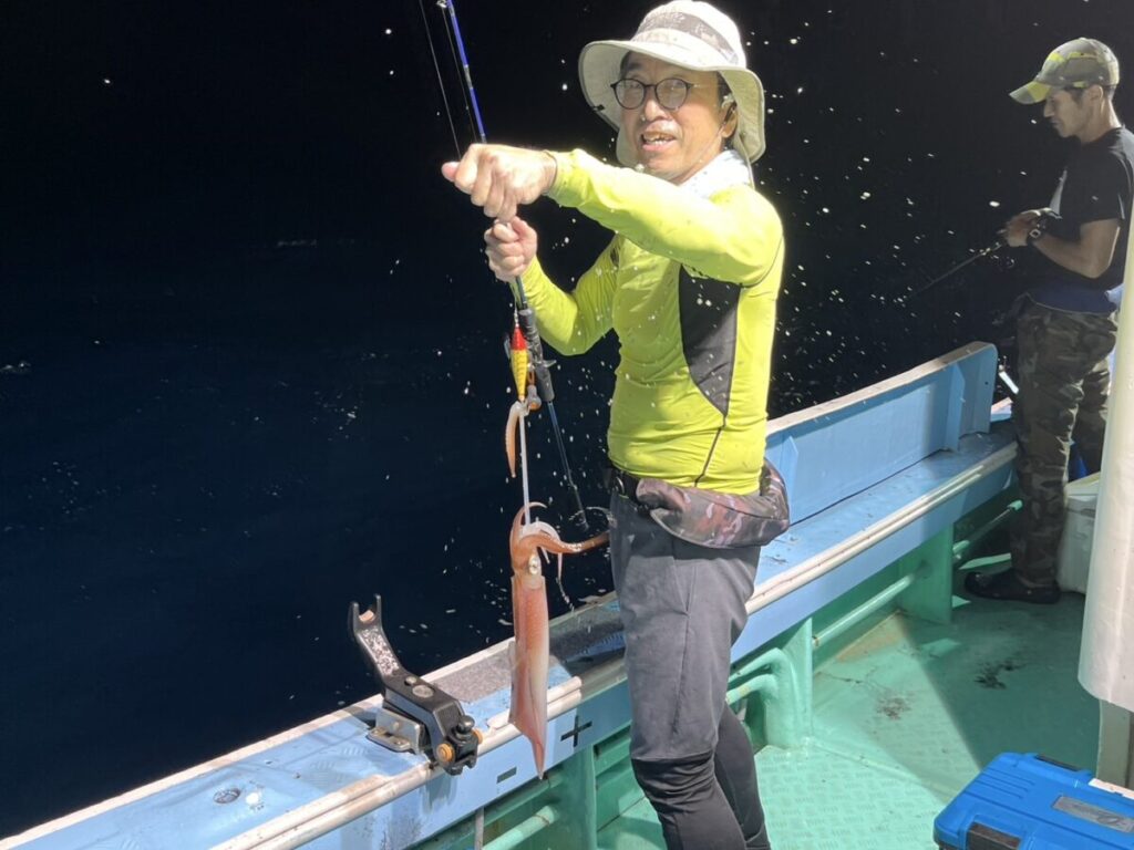 帽子に黄色のシャツを着た男が夜のボートの上に立ち、釣ったばかりのイカを載せた釣り竿を誇らしげに持っている。背後では迷彩柄のズボンをはいた別の人物が釣りをしている。
