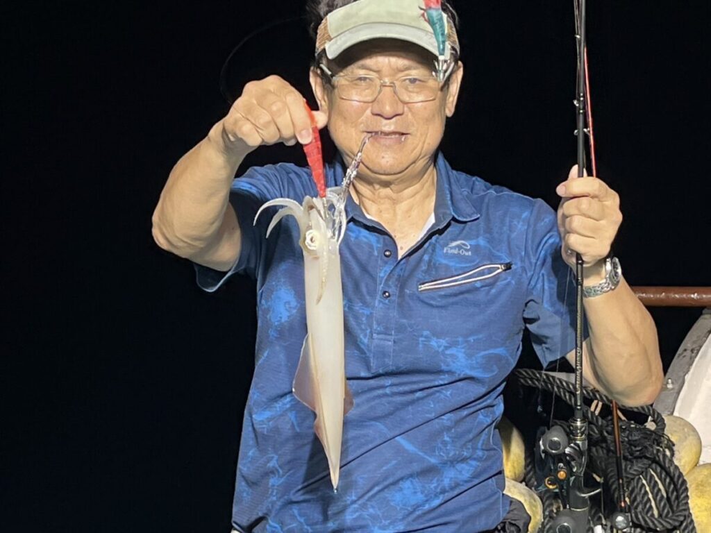 眼鏡をかけ、青いシャツを着た男が、片手に釣り竿、片手に釣りたてのイカを持ち、暗い背景の中で微笑んでいる。