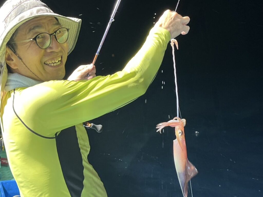 黄色いシャツに帽子をかぶった笑顔の人物が釣り竿を持ち、釣り糸には釣りたてのイカがぶら下がっている。背景は暗く、人物とイカを際立たせている。