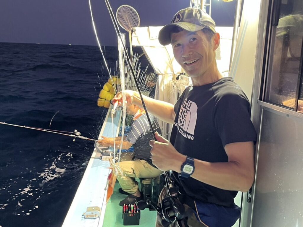 小魚と釣り竿を手に、釣りをしながら微笑む夜のボートの男。彼は帽子をかぶり、黒いTシャツを着て、腕時計をしている。背景には暗い海が見える。後ろにはもう一人座っている。