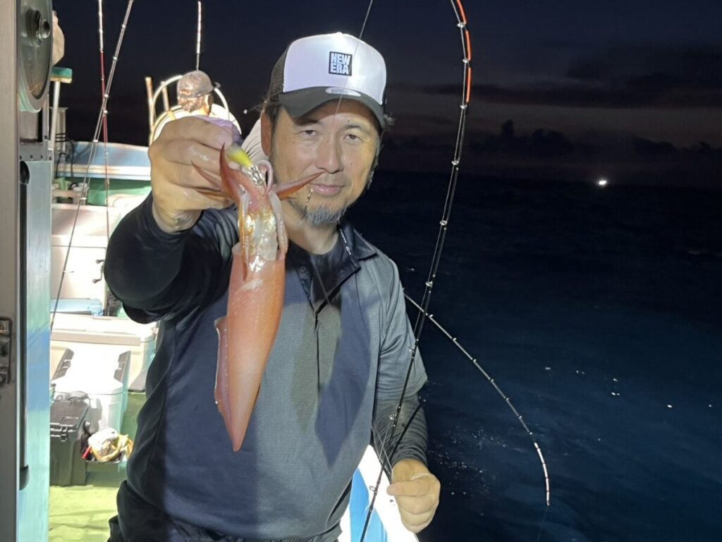 夜の船上で、釣り竿と暗い海水を背景に、釣りたてのイカを片手で掲げて微笑む男。