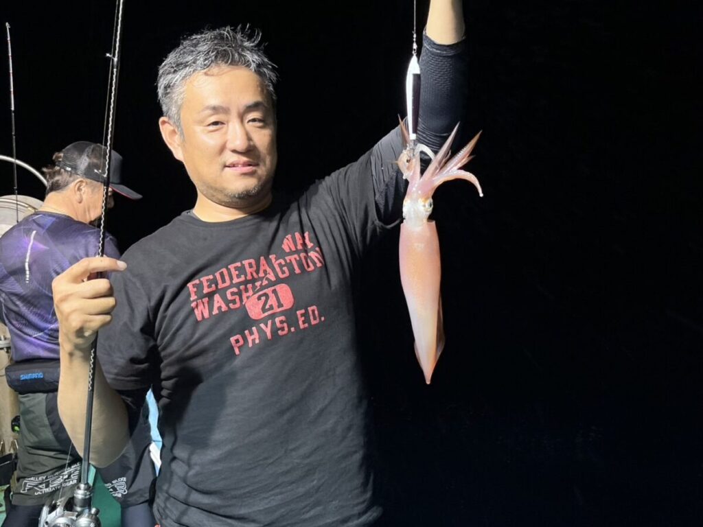 夜のボートに乗った男が、釣りたてのイカを誇らしげに片手で掲げ、その後ろで別の人が釣りをしている。.