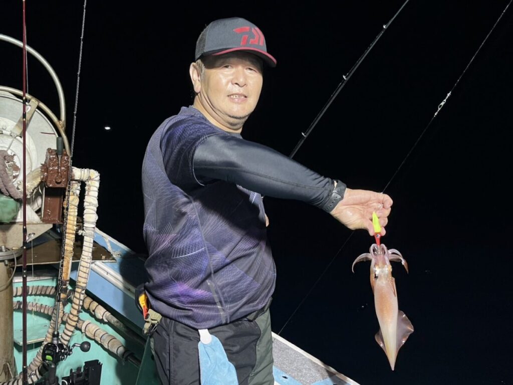 夜の船上で、釣り竿を持った釣りたてのイカを片手で持ち上げる人物。黒いシャツを着て、帽子をかぶり、カメラに向かって微笑んでいる。